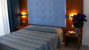 A hotel room with a large bed, a wooden headboard, two bedside lamps, a nightstand with a phone, and a vase with flowers. - Hotel Serena (Grado)