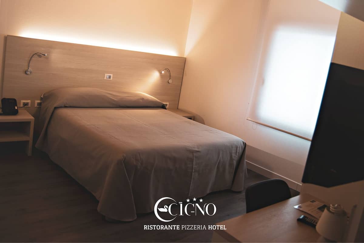 A hotel room with a bed, bedside table, lamp, TV, and a window.
