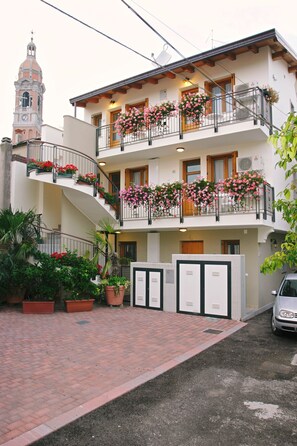 Um edifício de vários andares com varandas decoradas com flores, uma torre do relógio ao fundo e uma área de entrada pavimentada.