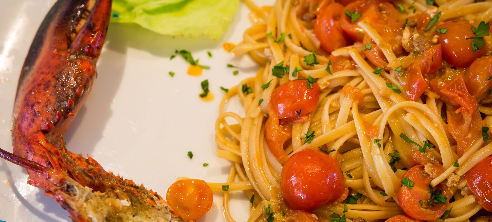 A plate of spaghetti with cherry tomatoes and parsley, and a cooked lobster on the side.