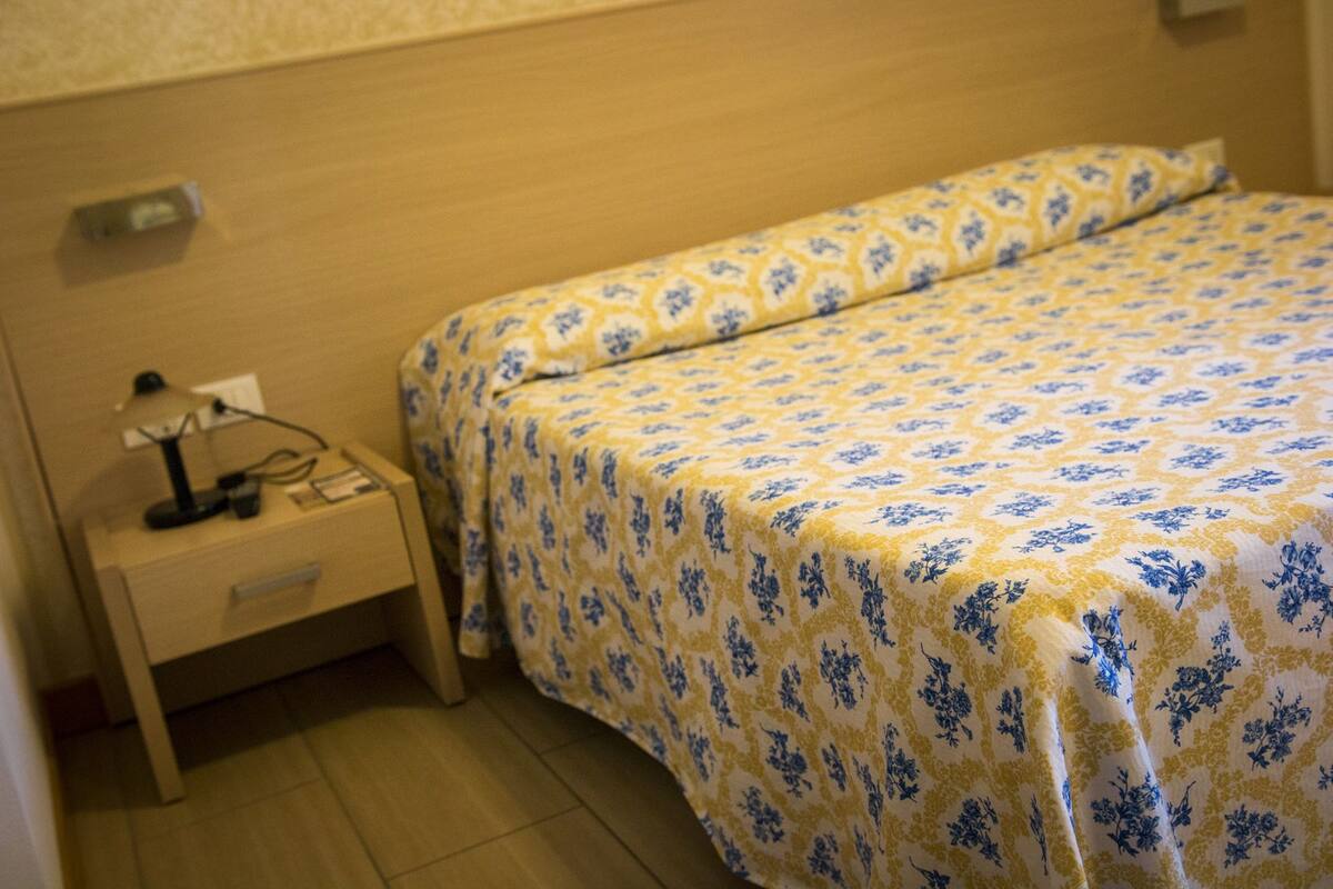 A hotel room with a bed covered in a floral-patterned bedspread, a bedside table with a lamp, and a power outlet.