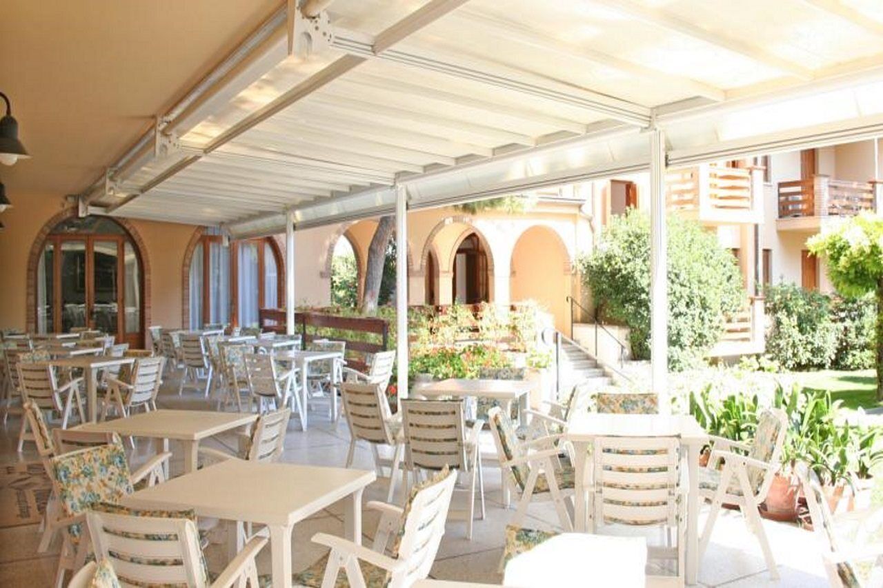 Um café ao ar livre com mesas e cadeiras brancas, coberto por um toldo branco, rodeado por vegetação e um edifício com varandas.