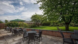 An outdoor patio area with round tables and chairs, a green umbrella, and a brick wall. - Woodcote Hotel (Ellesmere Port)