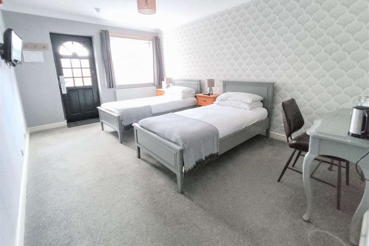 A hotel room with two single beds, a desk with a chair, a window with curtains, and a patterned wallpaper.