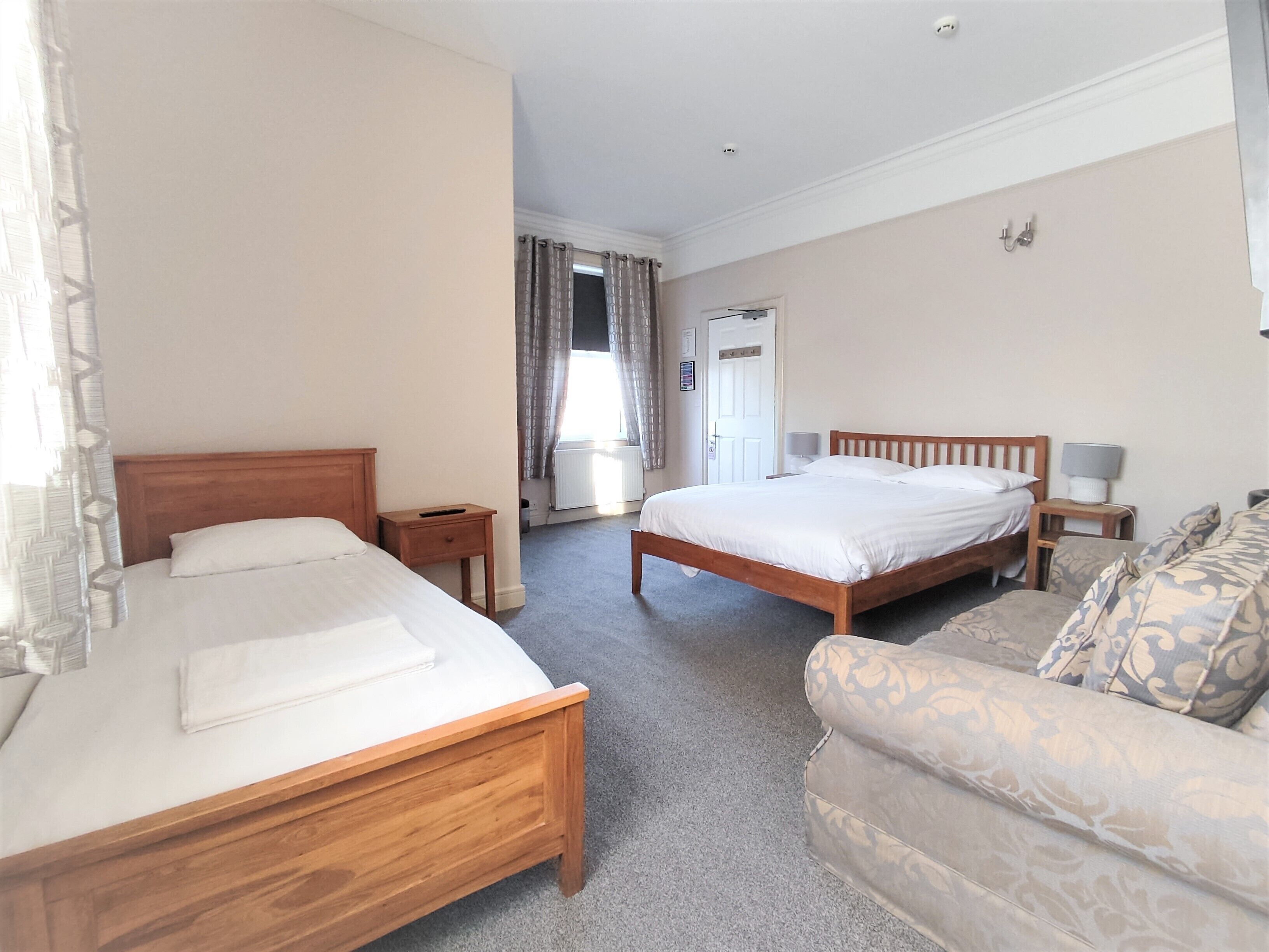 A bedroom with two wooden beds, a grey carpet, a patterned sofa, and a small bedside table with a lamp.