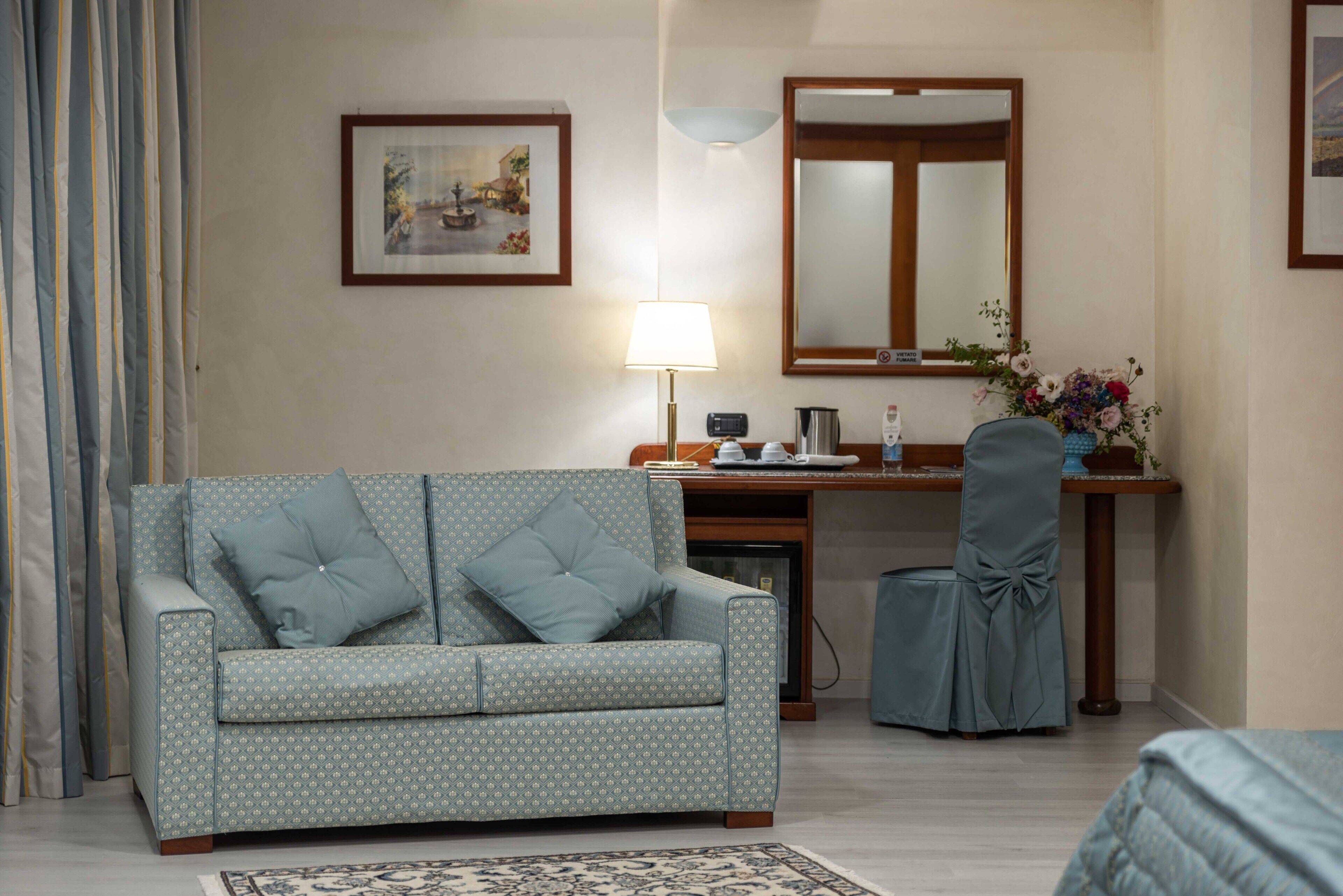 A hotel room with a patterned sofa, a desk with a lamp, a chair, a mirror, and a framed picture on the wall.