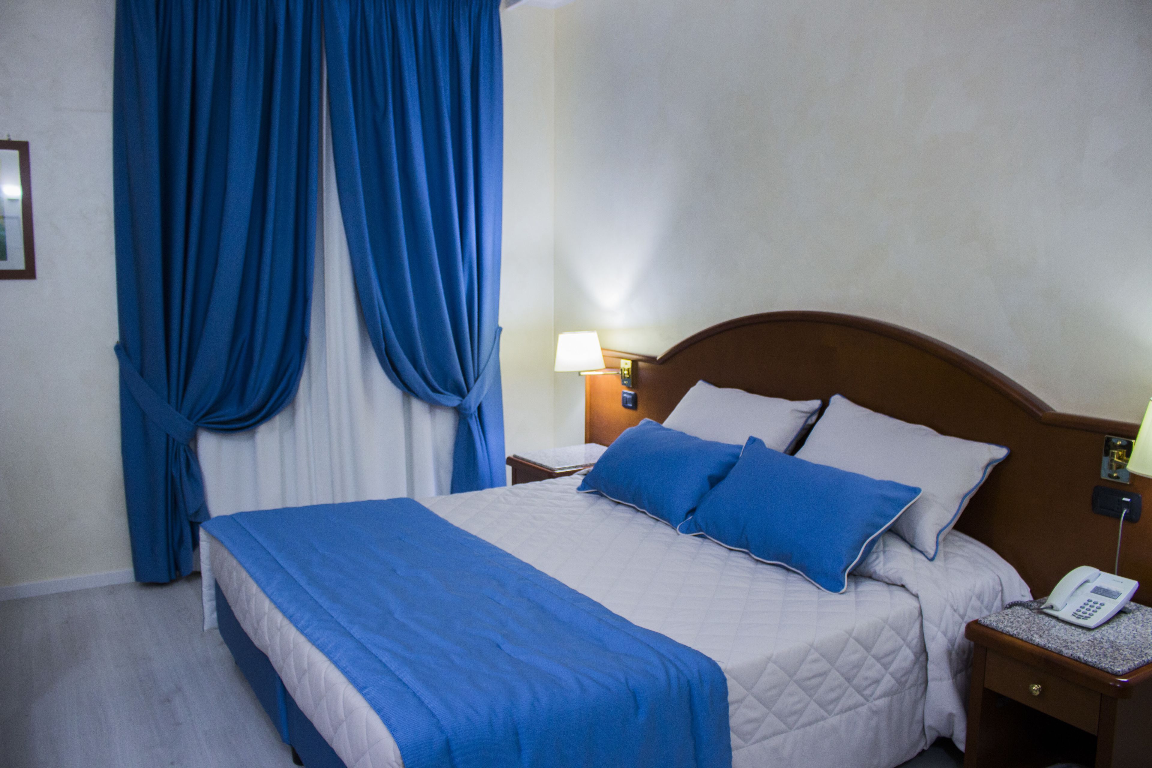 A hotel room with a bed, blue curtains, a bedside lamp, and a phone on the nightstand.