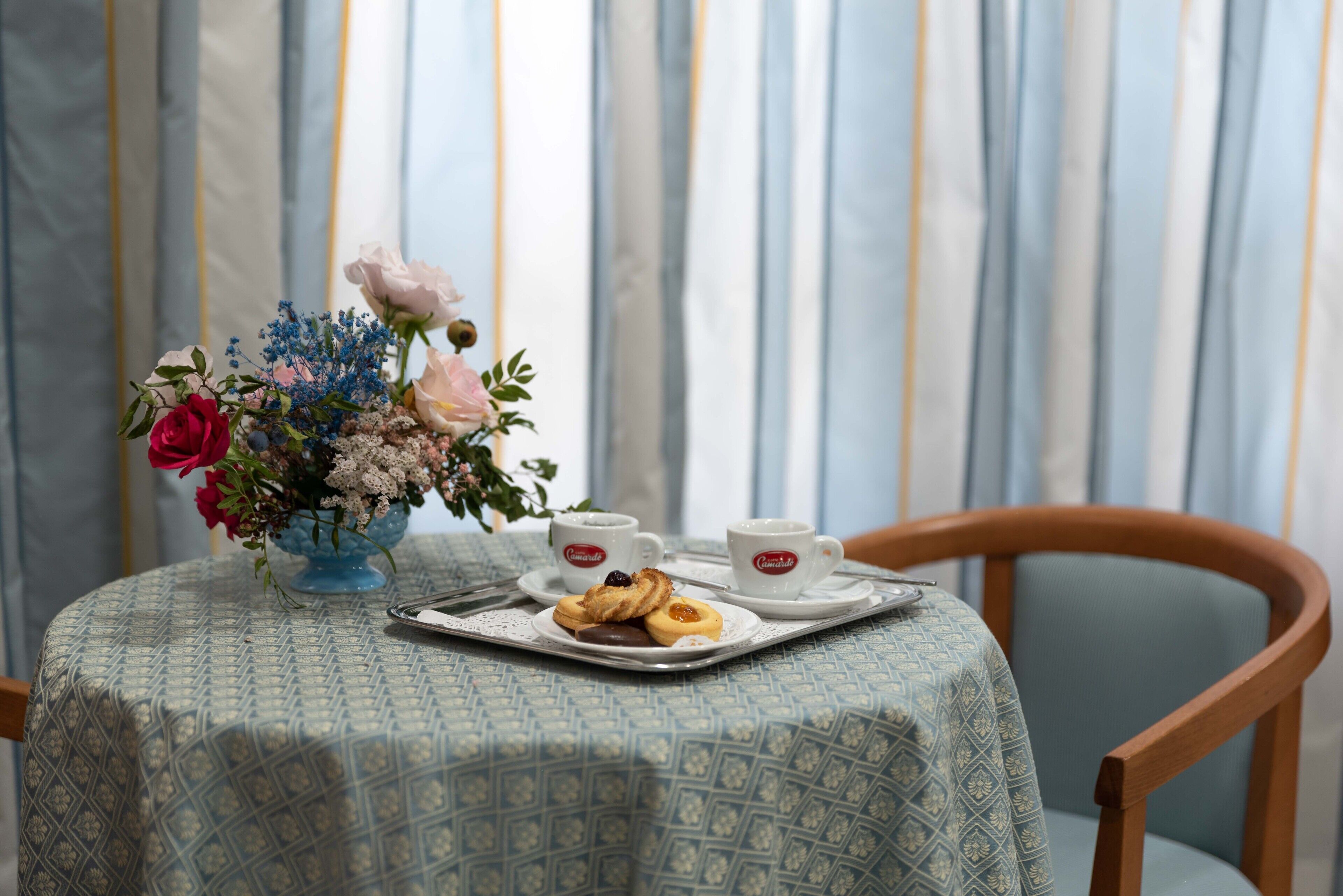 Een tafel met een tafelkleed met patroon, een bloemstuk, twee kopjes en een schaal met gebak.