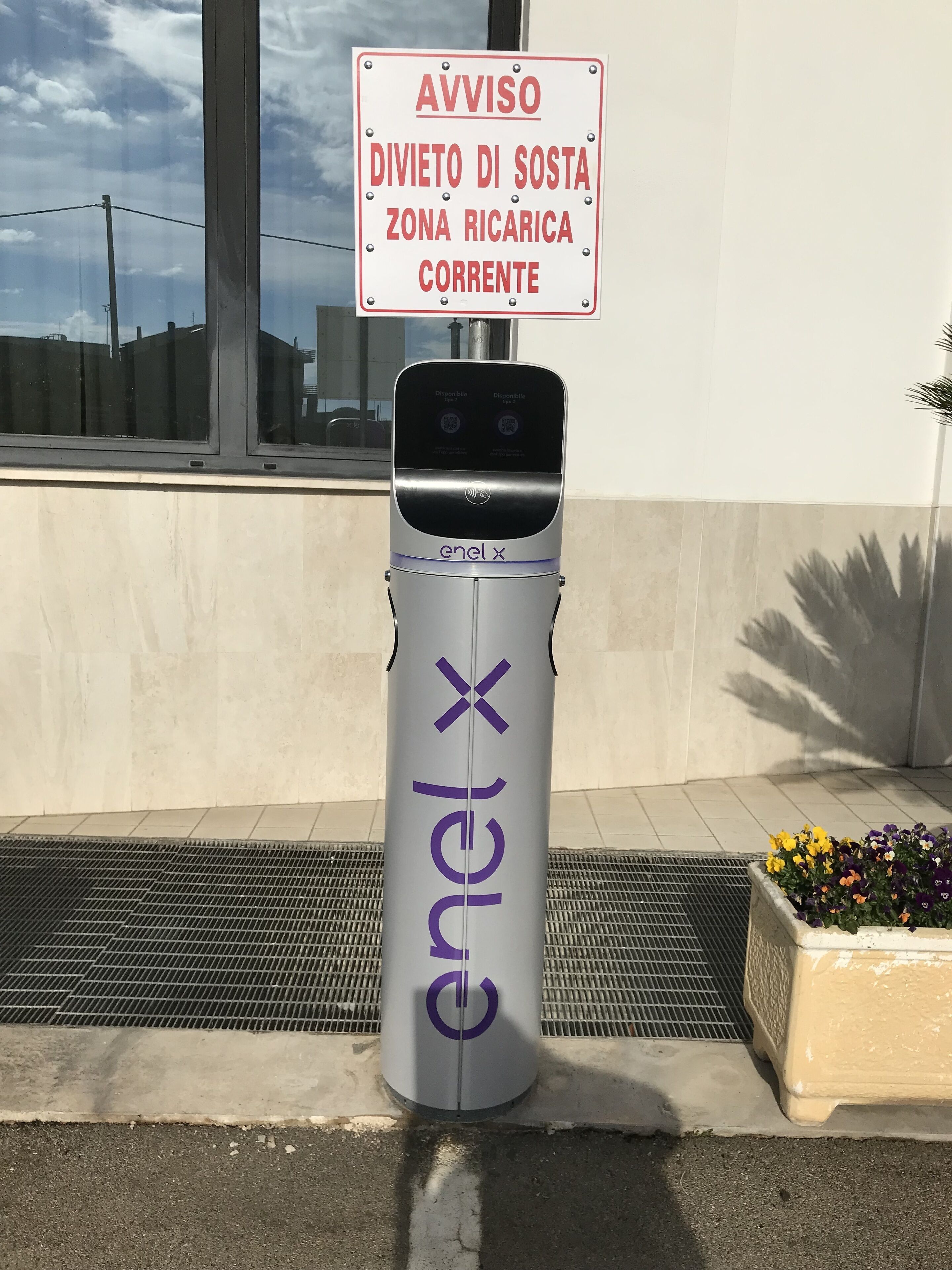 A public charging station branded 'enel X' with a sign above it warning of a 'Corrente' area.