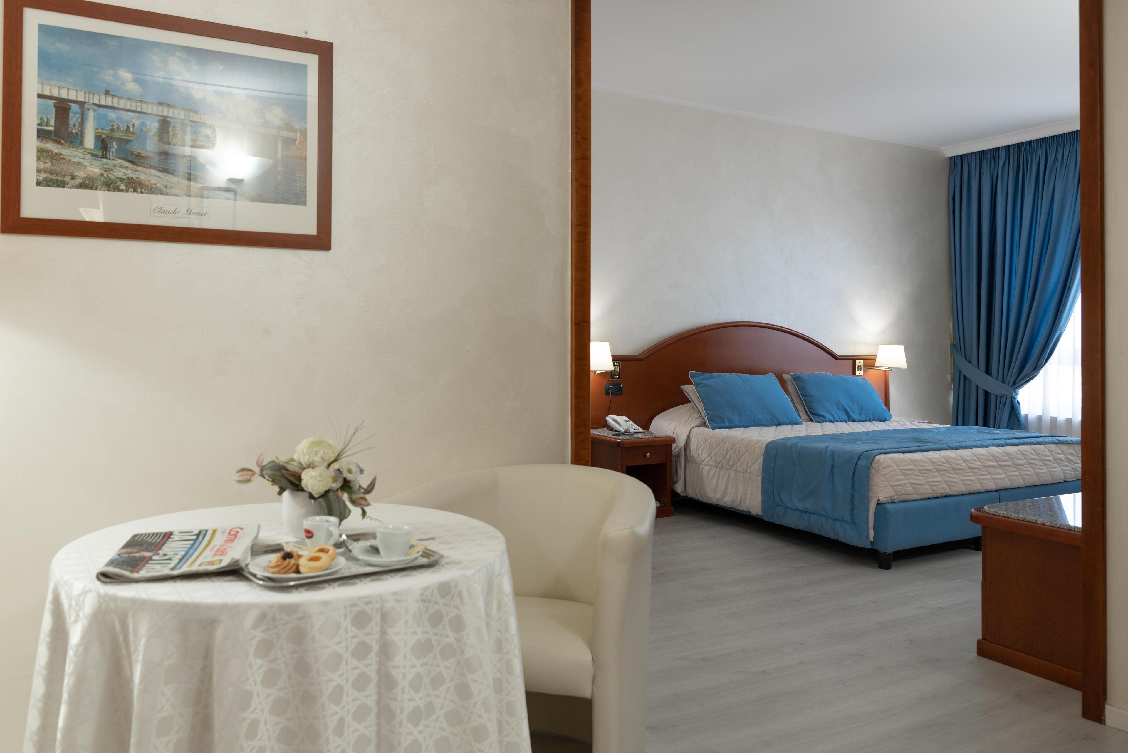 Habitación de hotel con cama, mesita de noche, una mesa con periódico y pasteles, y un cuadro en la pared.