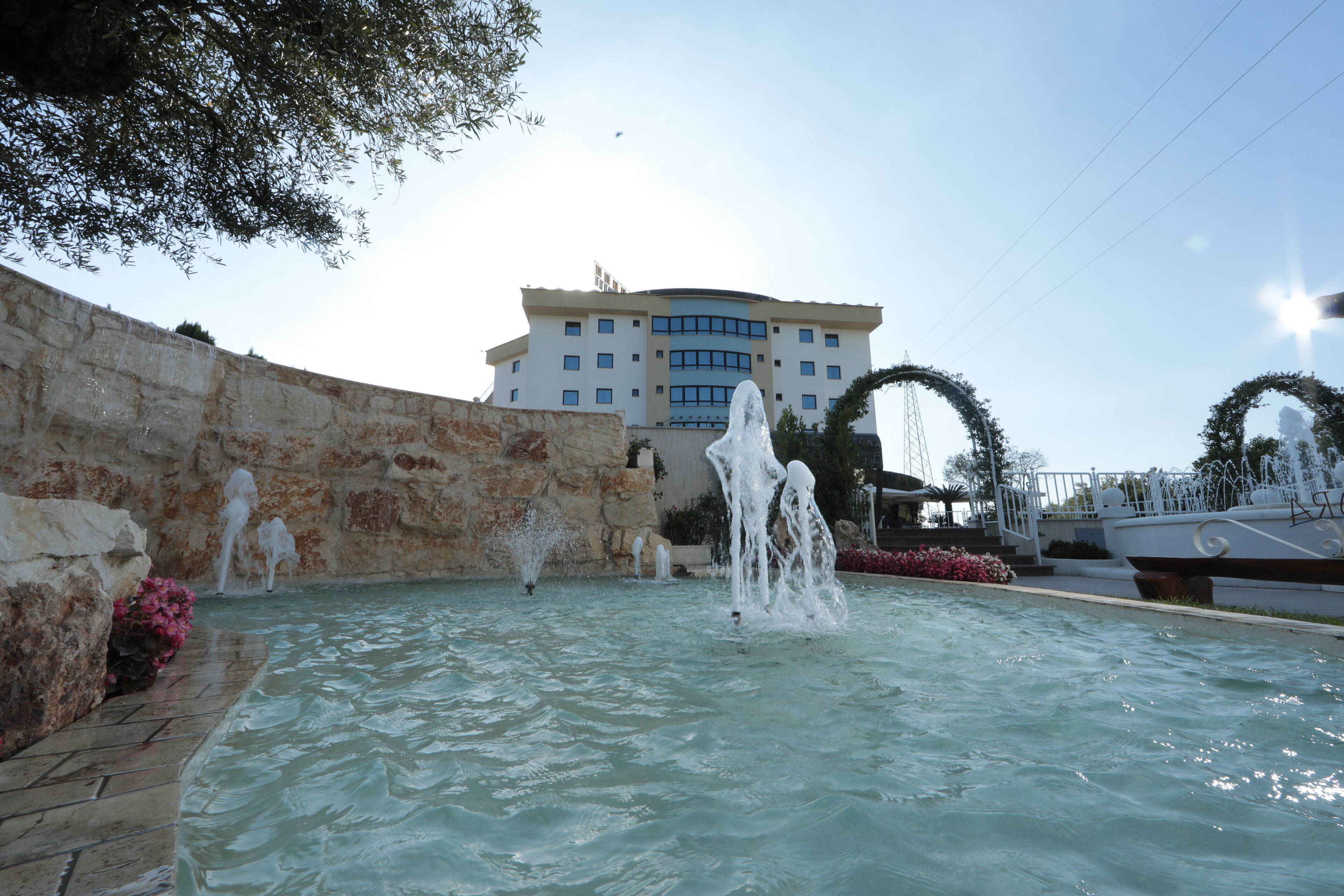 Una piscina con múltiples chorros de agua, un muro de piedra y un edificio con techo curvo al fondo.