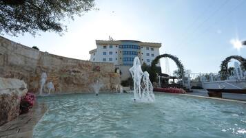Una piscina con múltiples chorros de agua, un muro de piedra y un edificio con techo curvo al fondo.
