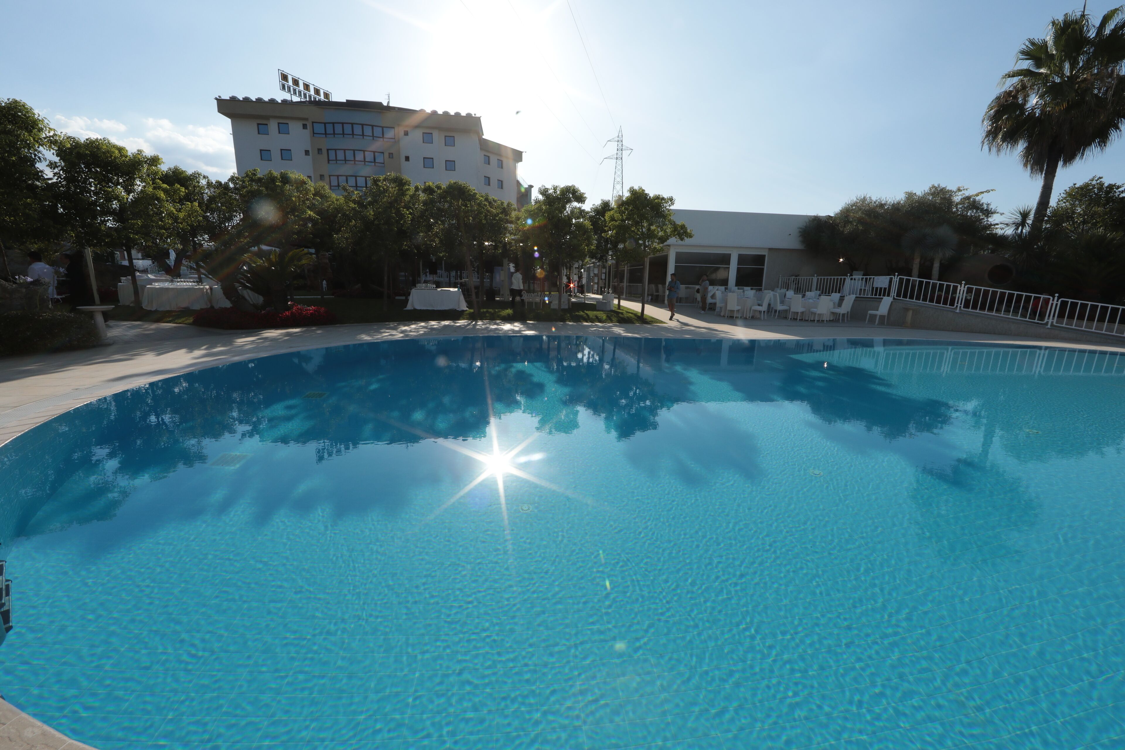 A clear blue pool with a sun reflection, surrounded by trees and a building in the background.