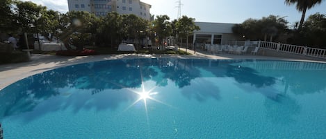 A clear blue pool with a sun reflection, surrounded by trees and a building in the background.