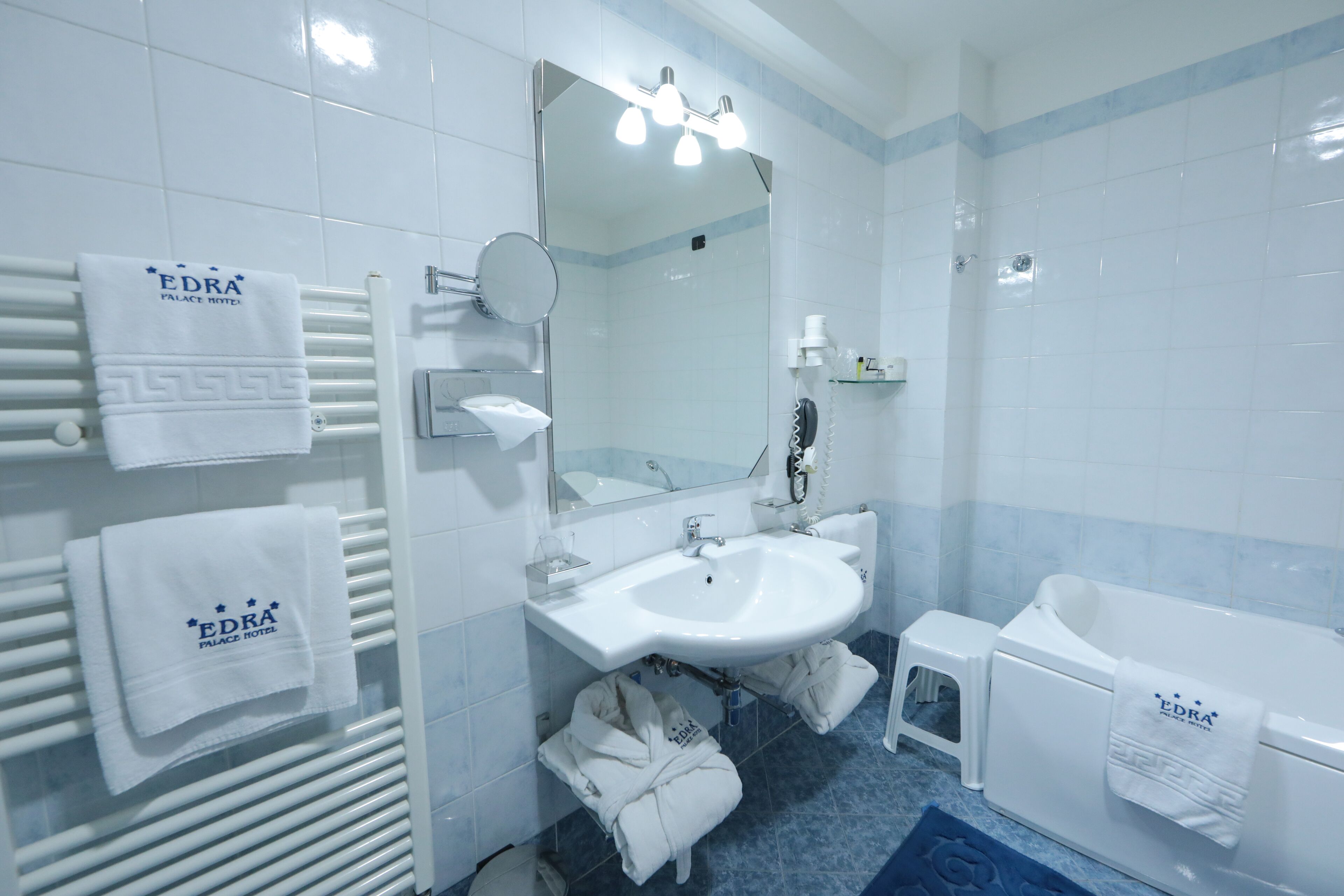A modern bathroom with a white sink, mirror, and bathtub. There are towels hanging on the wall and a small stool near the bathtub.
