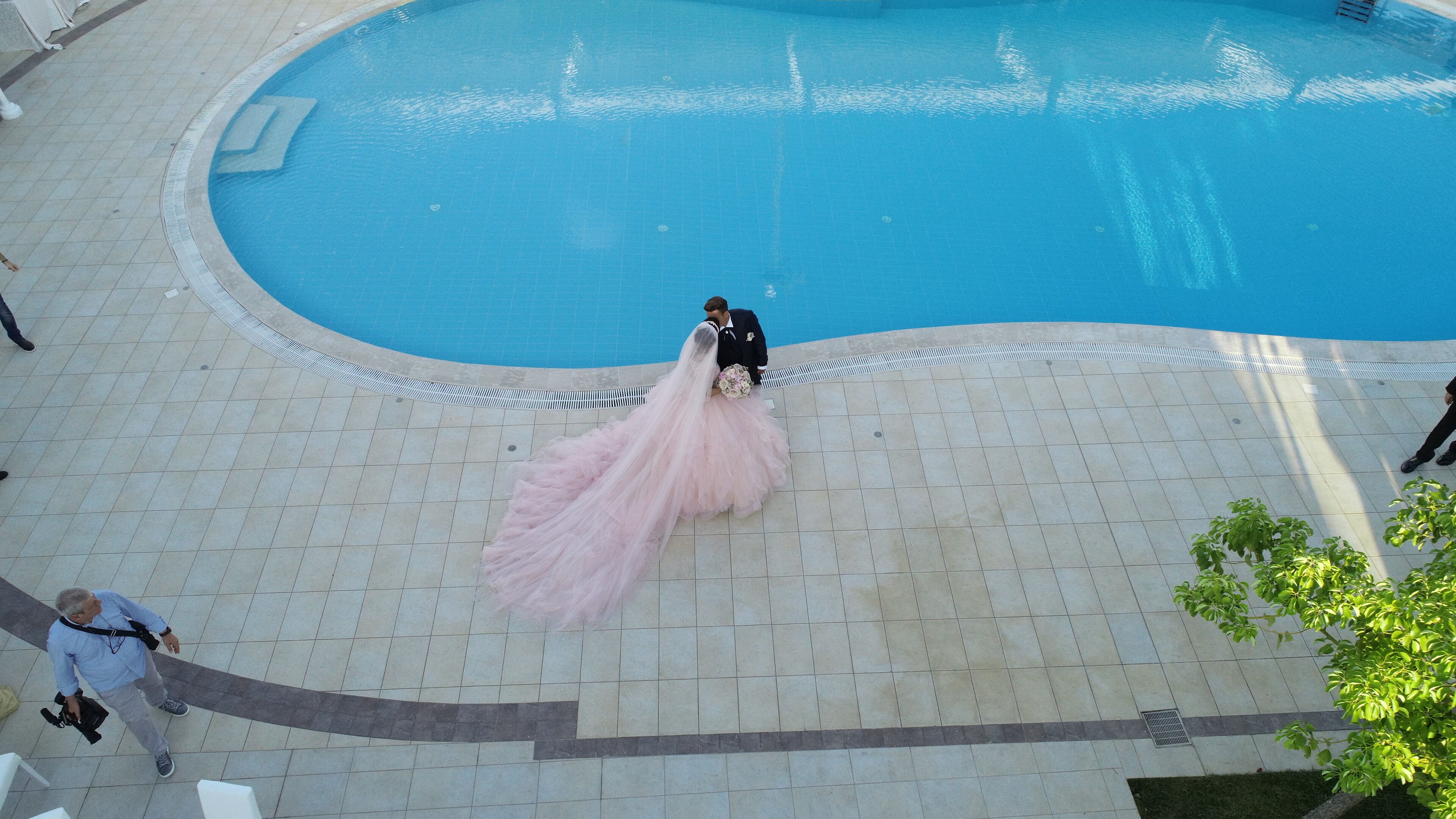 A wedding scene with a bride in a pink dress and groom in a black suit by a pool.