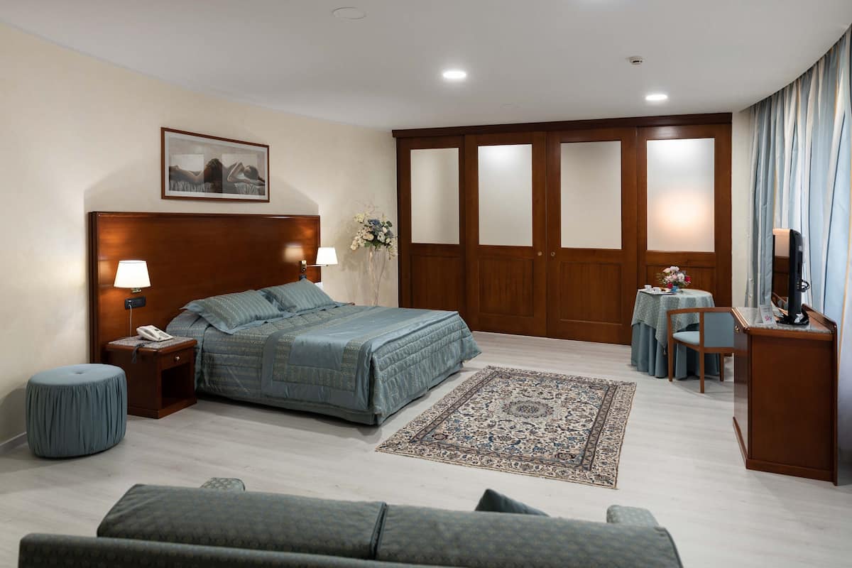 A bedroom with a large bed, a nightstand with a lamp, a small table with a vase of flowers, a wooden wardrobe, and a television on a stand.