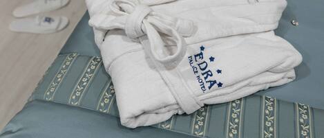 Un albornoz blanco impecablemente doblado con el bordado "EDRA PALACE HOTEL", sobre un cojín azul con estampado floral.
