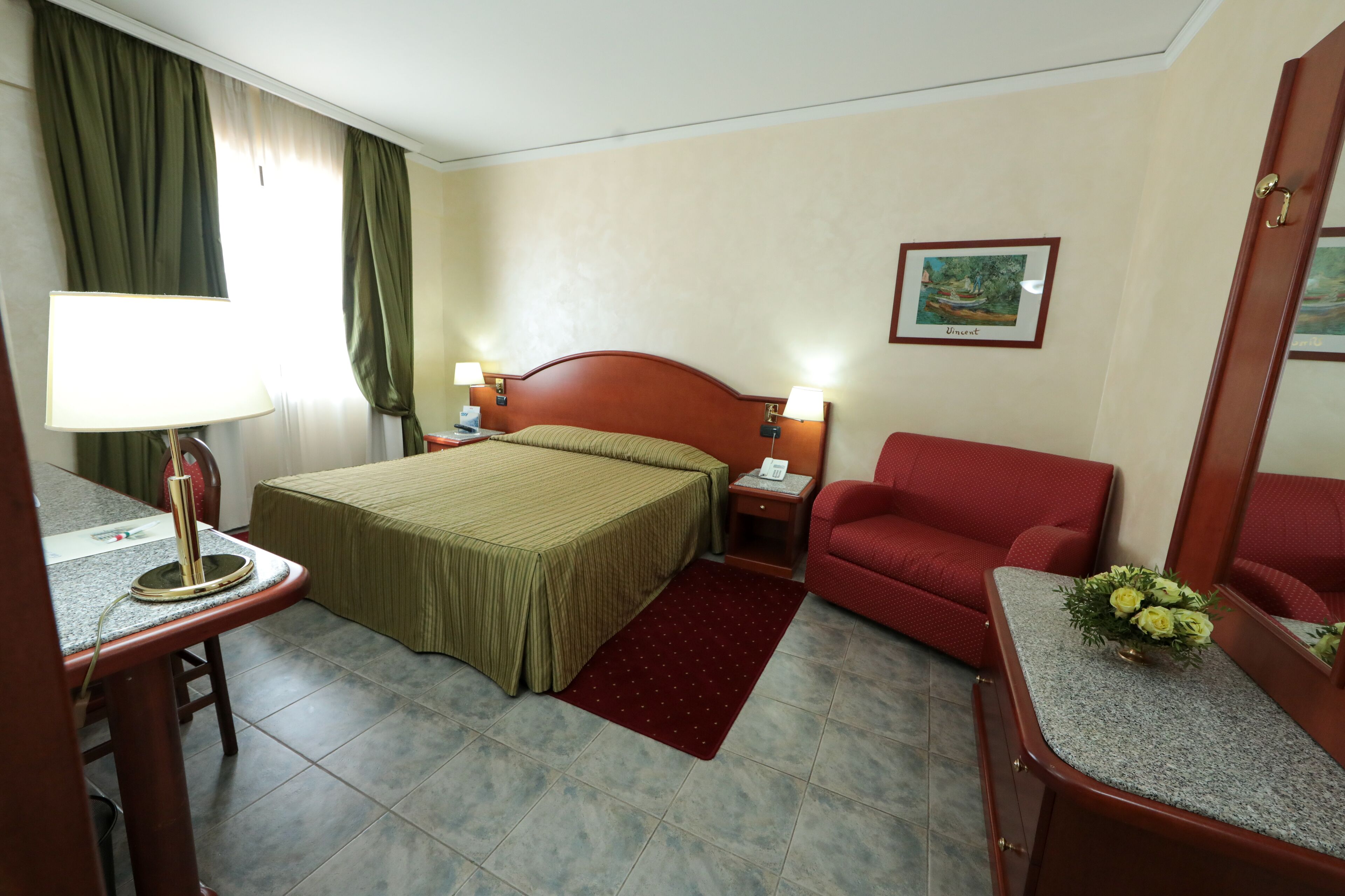 A hotel room with a bed, two bedside lamps, a red armchair, a small table with a lamp, a red rug, and a framed picture on the wall.
