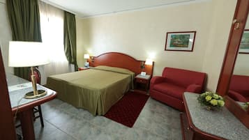 A hotel room with a bed, two bedside lamps, a red armchair, a small table with a lamp, a red rug, and a framed picture on the wall.