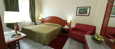 Habitación de hotel con una cama, dos lámparas de noche, un sillón rojo, una mesita con lámpara, una alfombra roja y un cuadro enmarcado en la pared.