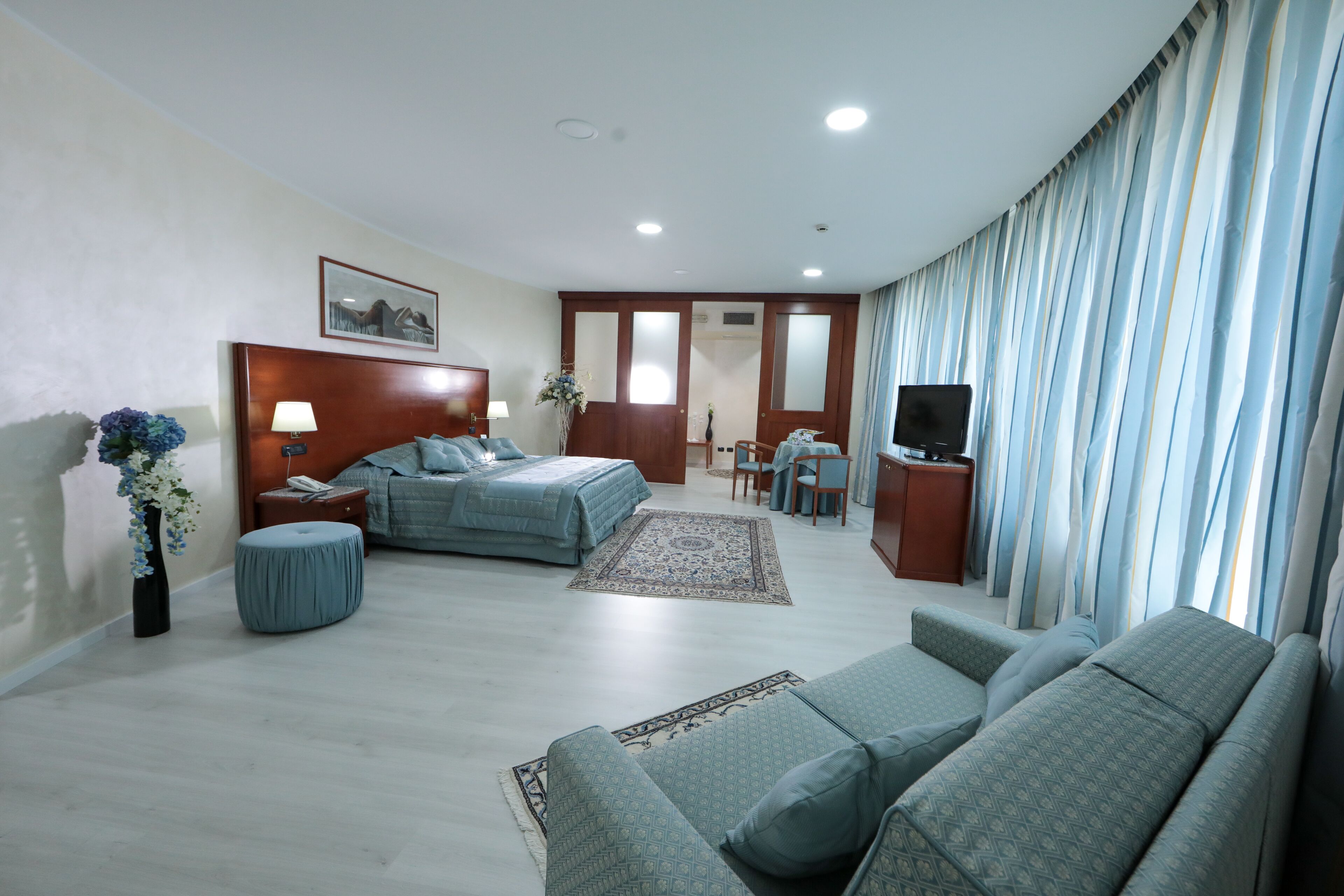 A hotel room with a bed, a sofa, a desk, a TV, and a large window with curtains.