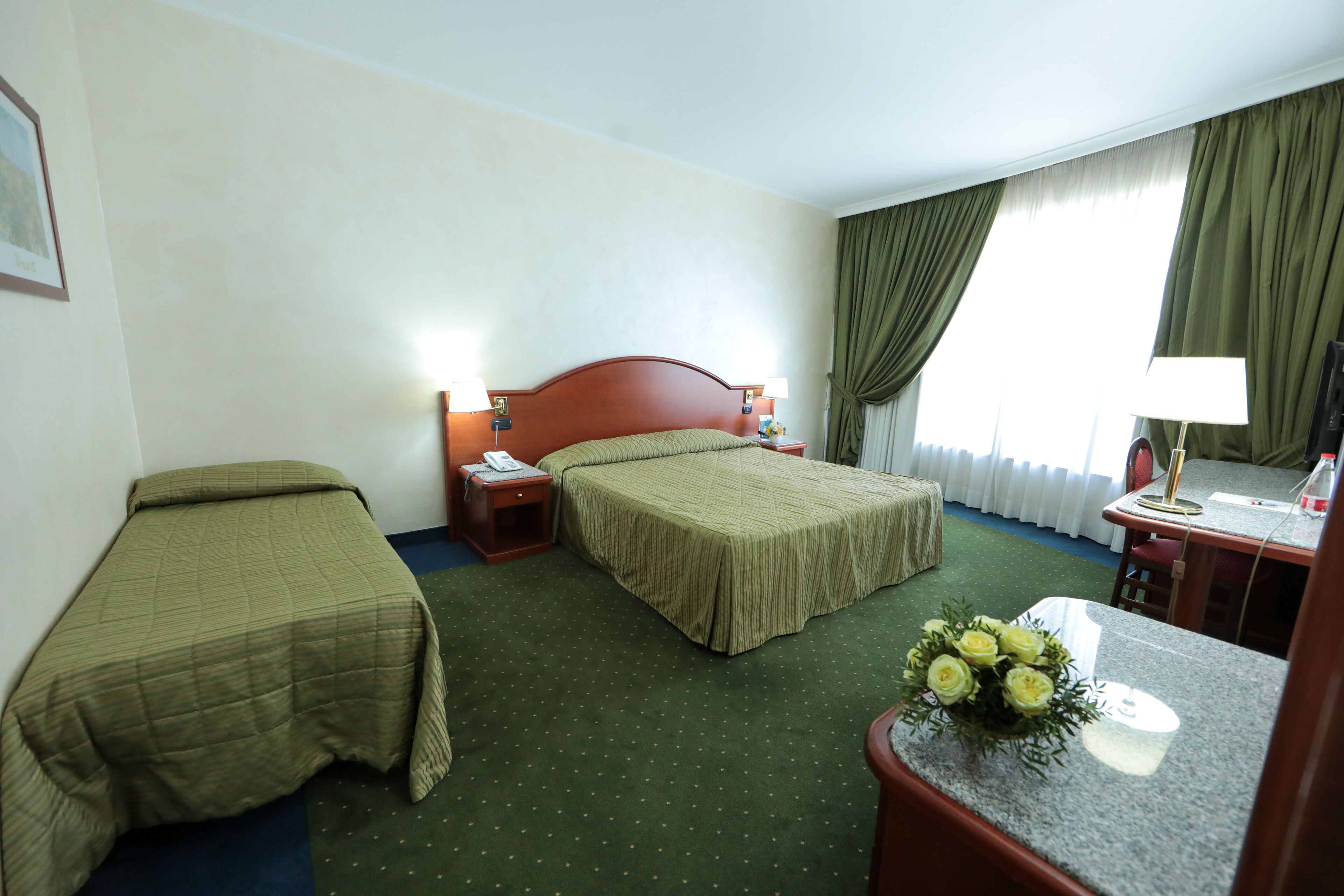Habitación de hotel con dos camas individuales, un escritorio con silla, una mesita de noche con lámpara y un ramo de flores.