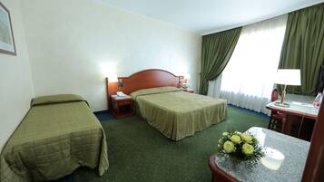 Habitación de hotel con dos camas individuales, un escritorio con silla, una mesita de noche con lámpara y un ramo de flores.