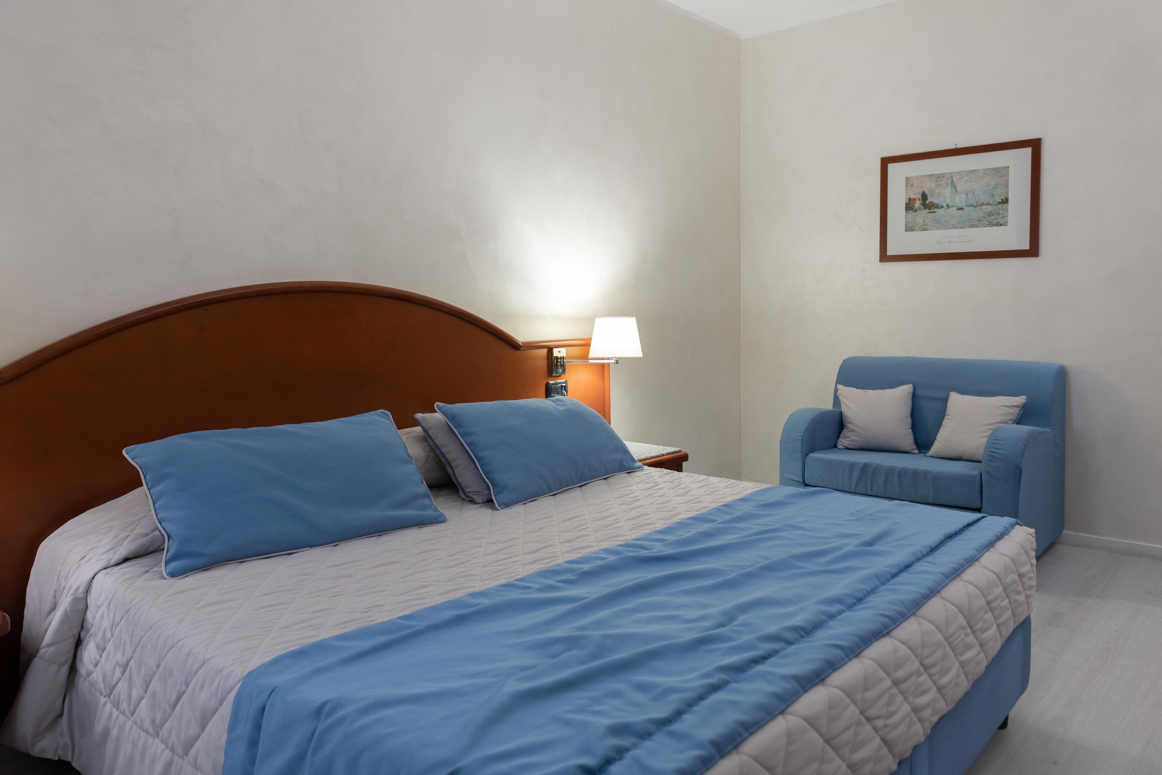 Un lit bien fait, avec une literie bleue, une tête de lit en bois, une lampe de chevet et un fauteuil bleu agrémenté d’un coussin.