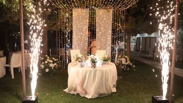 Decoración para una boda al aire libre, con una mesa cubierta por un mantel blanco, sillas a su alrededor y adornos de flores y velas.
