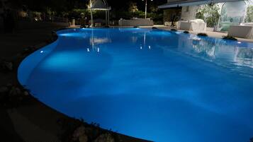 Ein Pool bei Nacht mit geschwungener Kante, umgeben von weißen Liegestühlen und einer überdachten Sitzgruppe.