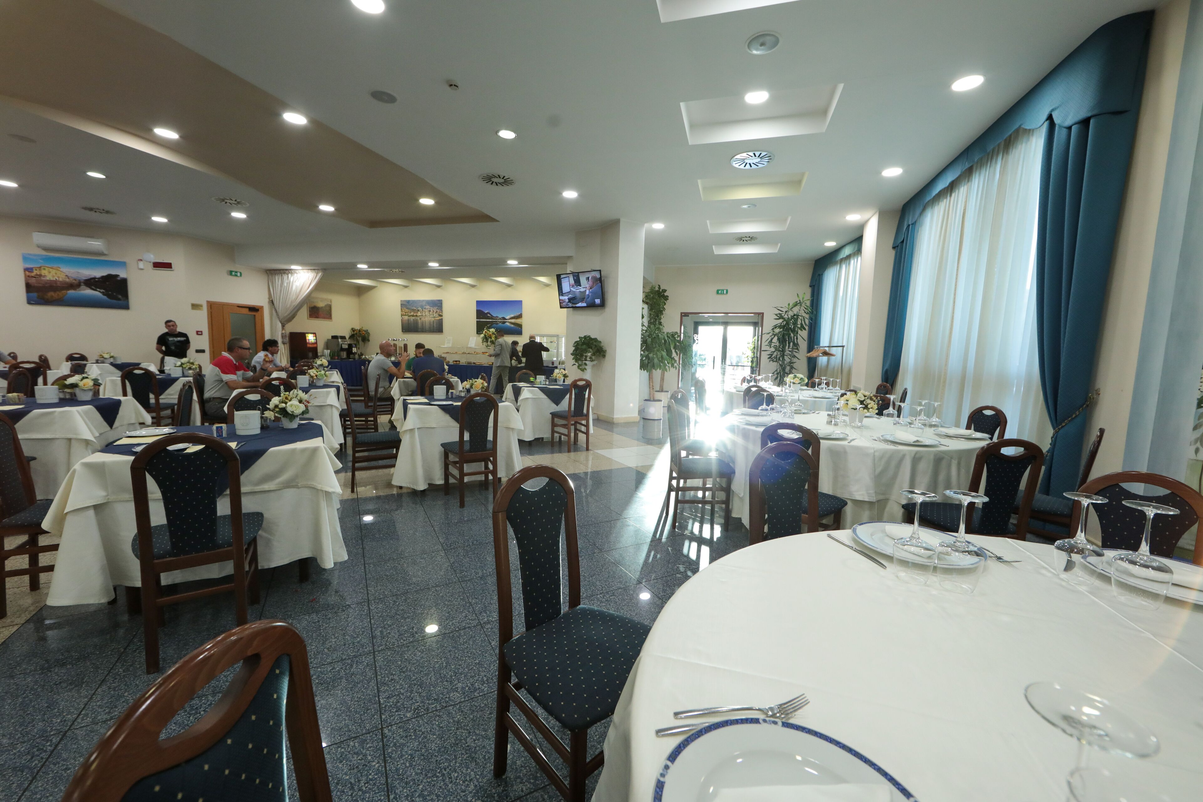 A spacious dining area with multiple tables set for guests, a television mounted on the wall, and a large window with curtains.