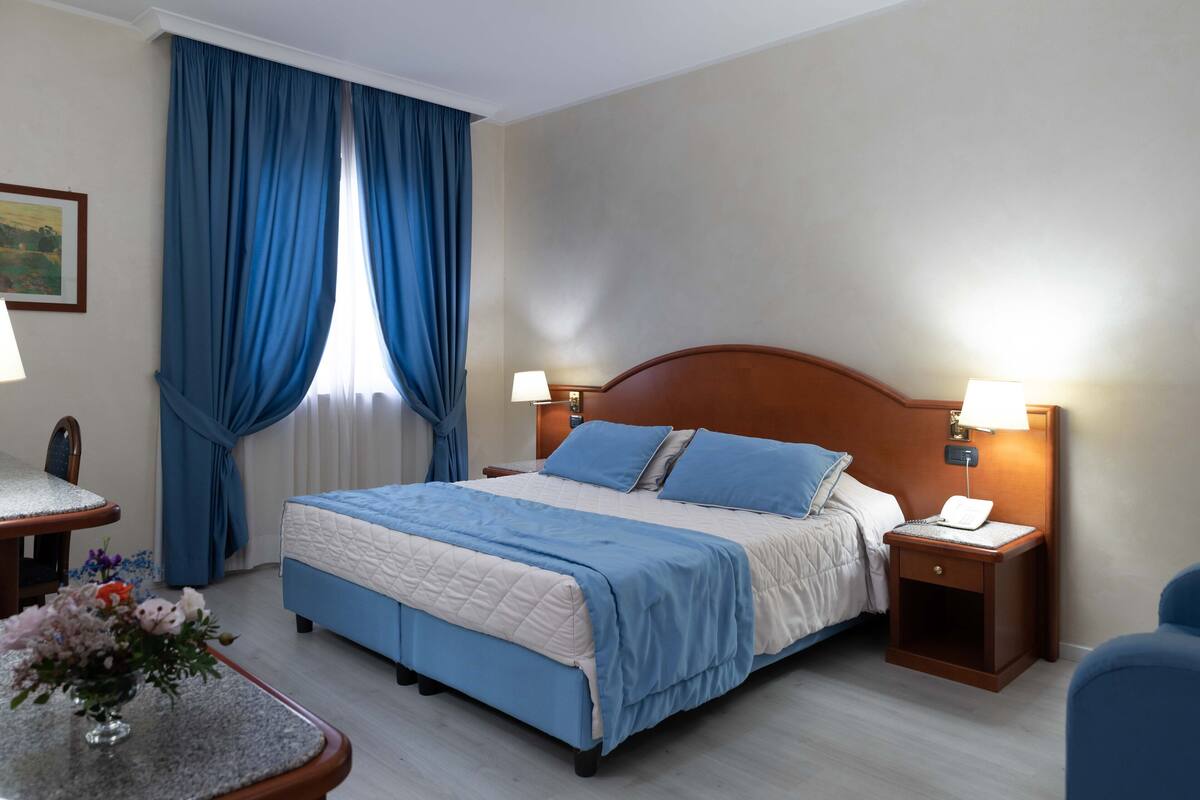 A hotel room with a bed, two bedside tables, a chair, a desk, and a window with blue curtains.