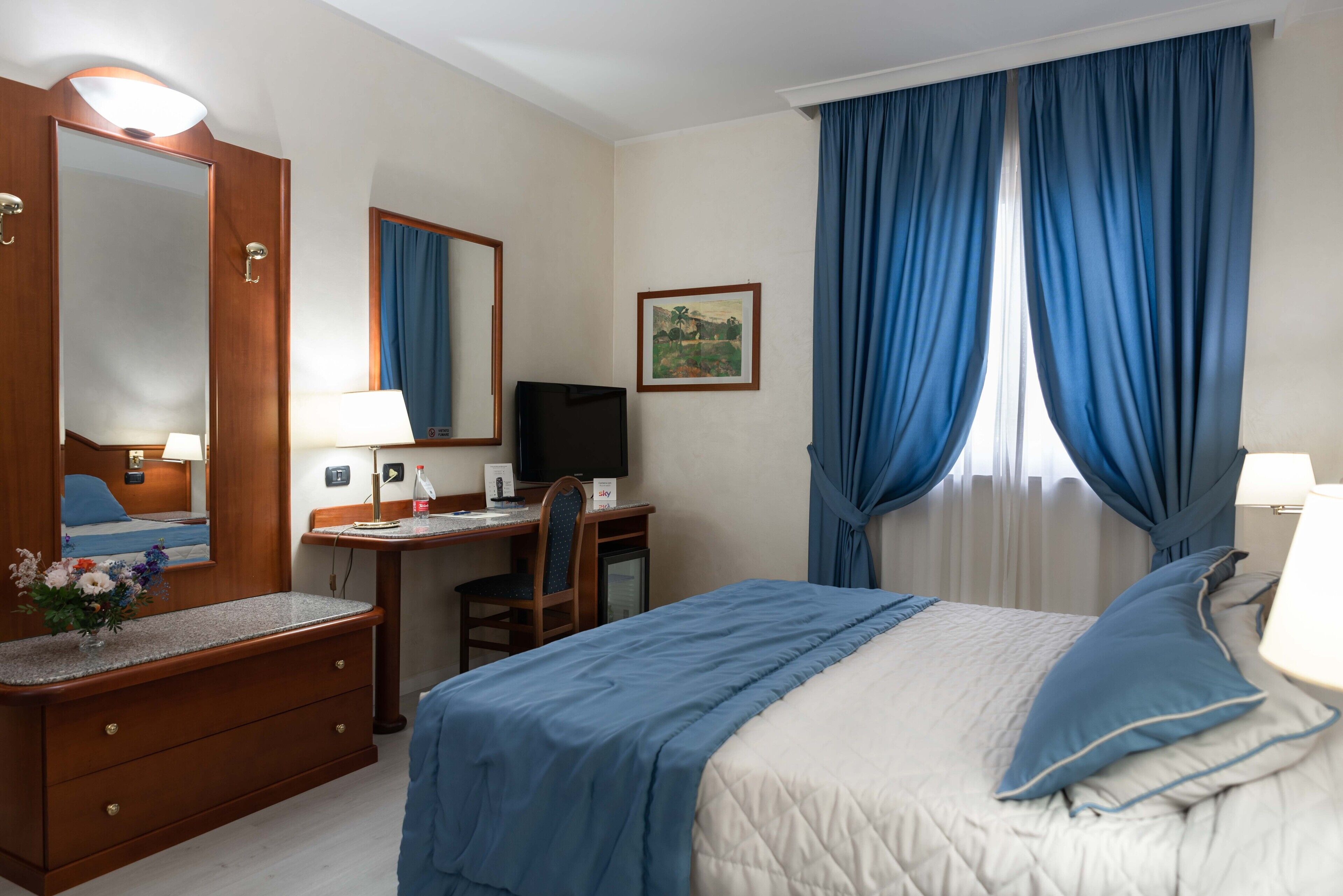 A hotel room with a bed, a desk with a computer, a TV, a mirror, and a window with blue curtains.