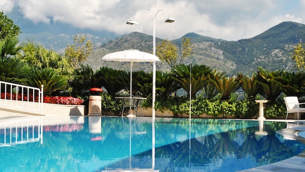 A swimming pool with a white umbrella, surrounded by lush greenery and mountains in the background. - Edra Palace (Cassino)