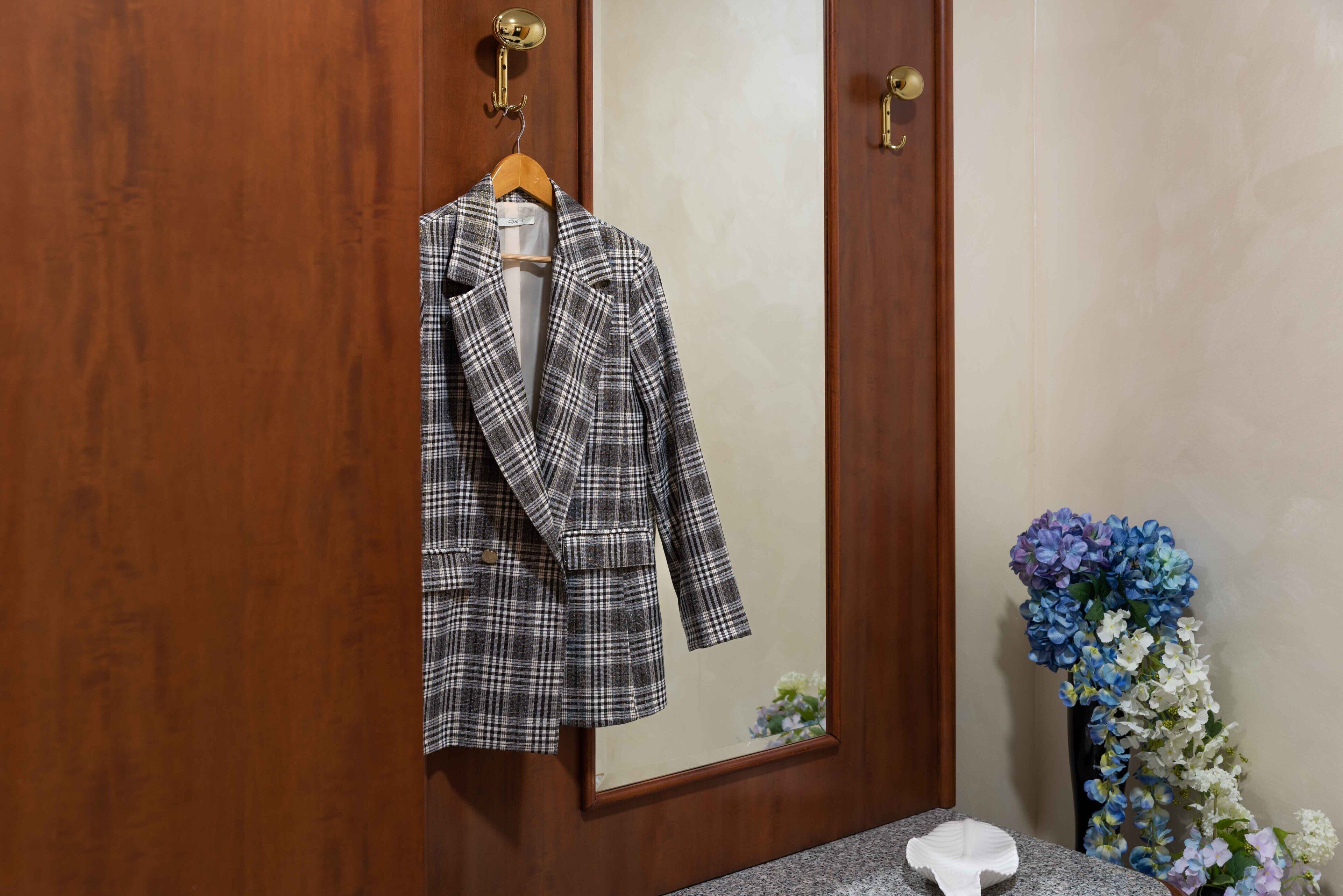 Un blazer escocés colgado en una puerta de madera, un espejo y un jarrón con flores.
