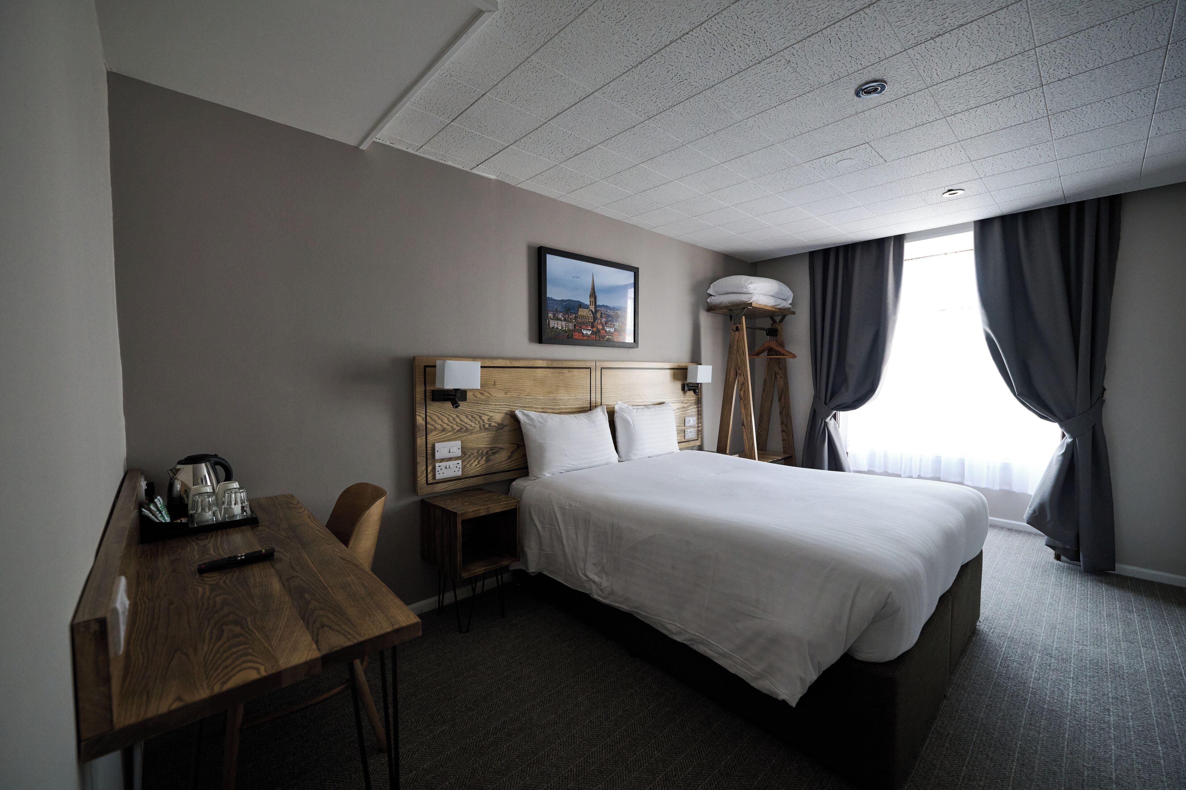 A hotel room with a bed, a desk with a kettle, a chair, and a picture on the wall.
