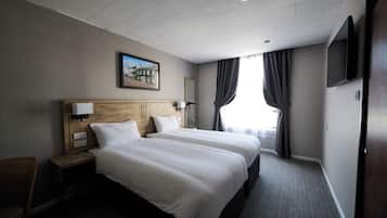 A hotel room with two beds, a wooden headboard, a nightstand with a lamp, a TV mounted on the wall, and a window with curtains.
