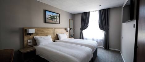 Habitación de hotel con dos camas, cabecero de madera, mesita de noche con lámpara, televisión montada en la pared y ventana con cortinas.