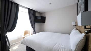 A hotel room with a large bed, a flat-screen TV mounted on the wall, a wooden headboard, and a window with curtains.