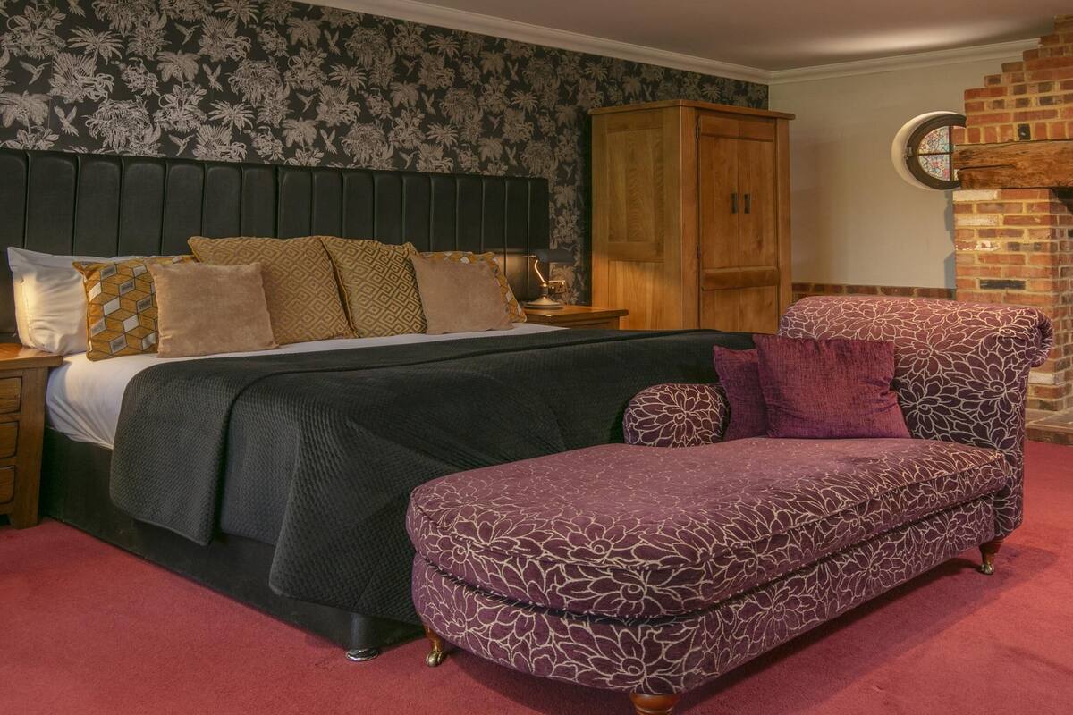 A bedroom with a bed, a wooden wardrobe, a brick fireplace, and a patterned armchair.