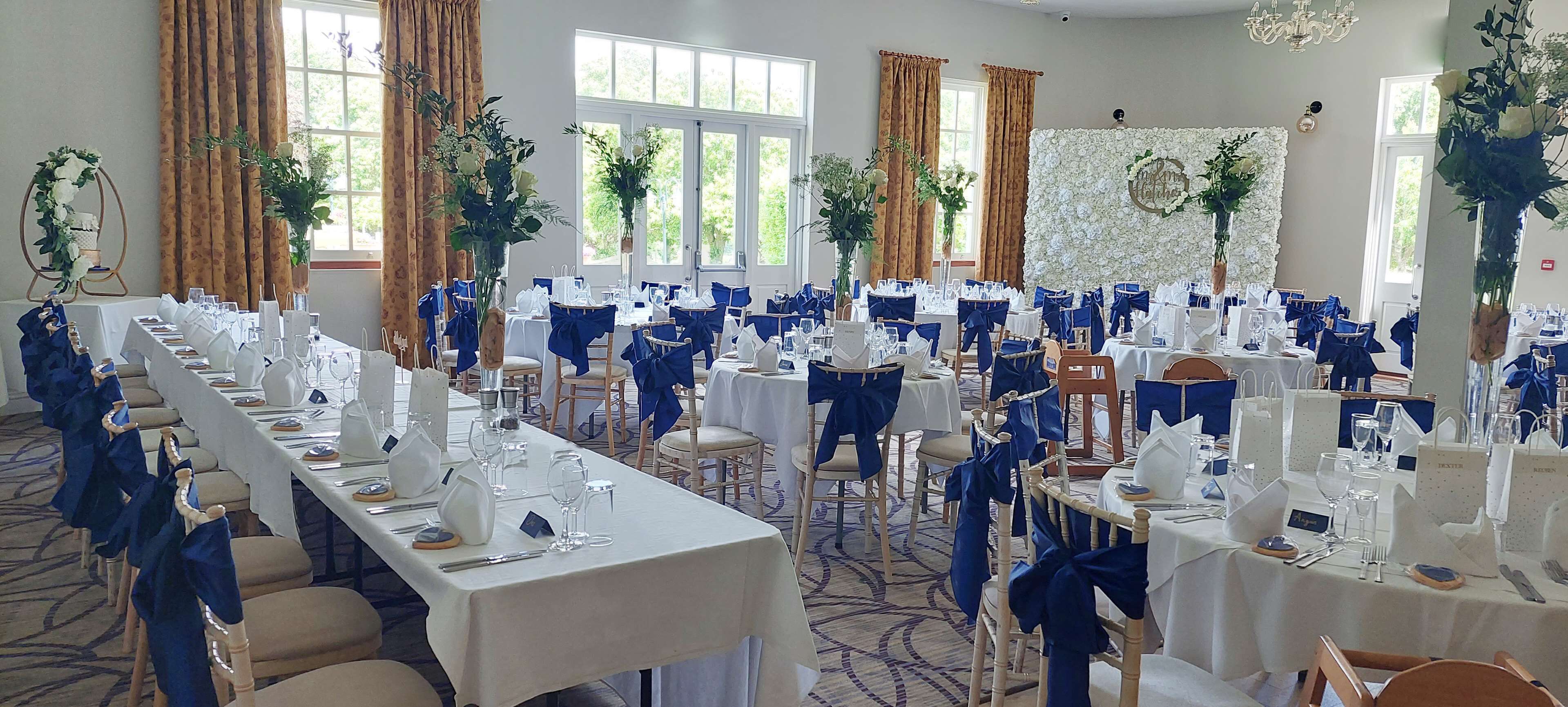 Una sala de banquets amb taules llargues preparades per a un esdeveniment formal, decorada amb fundes blaves per als seients i tovallons blancs.
