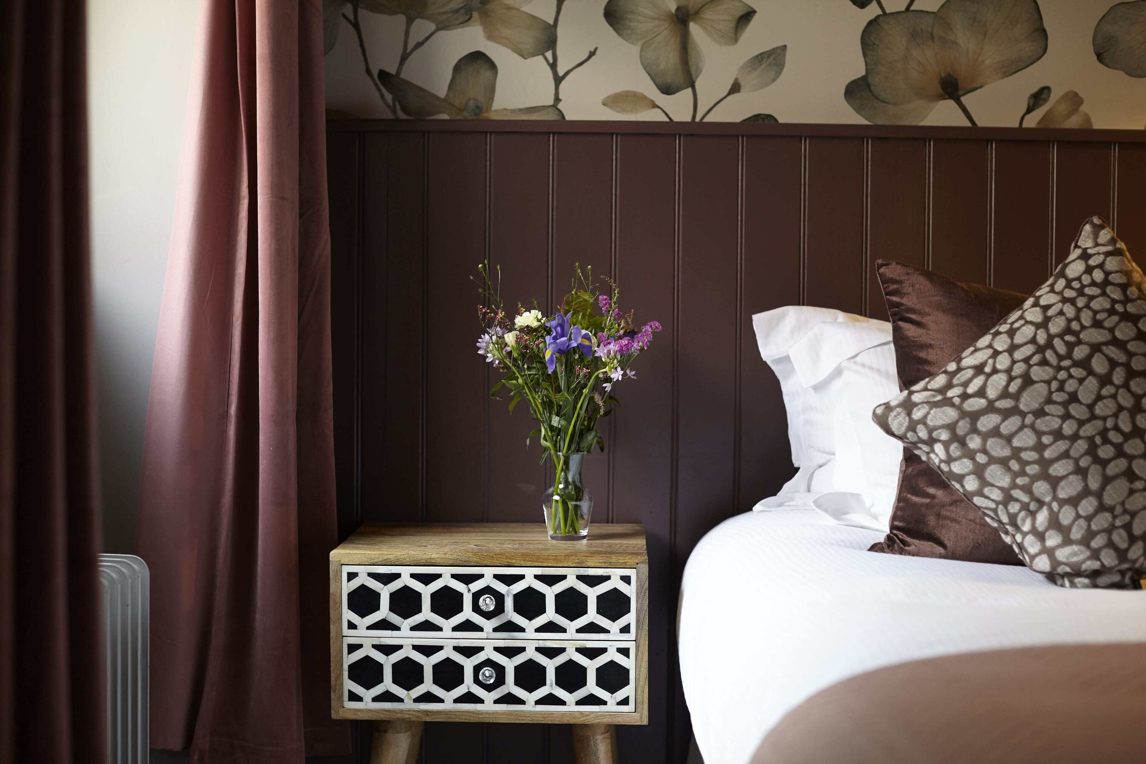 Una cama bien hecha con un cojín estampado, una mesita de noche con un jarrón de flores y una pared con un diseño floral.