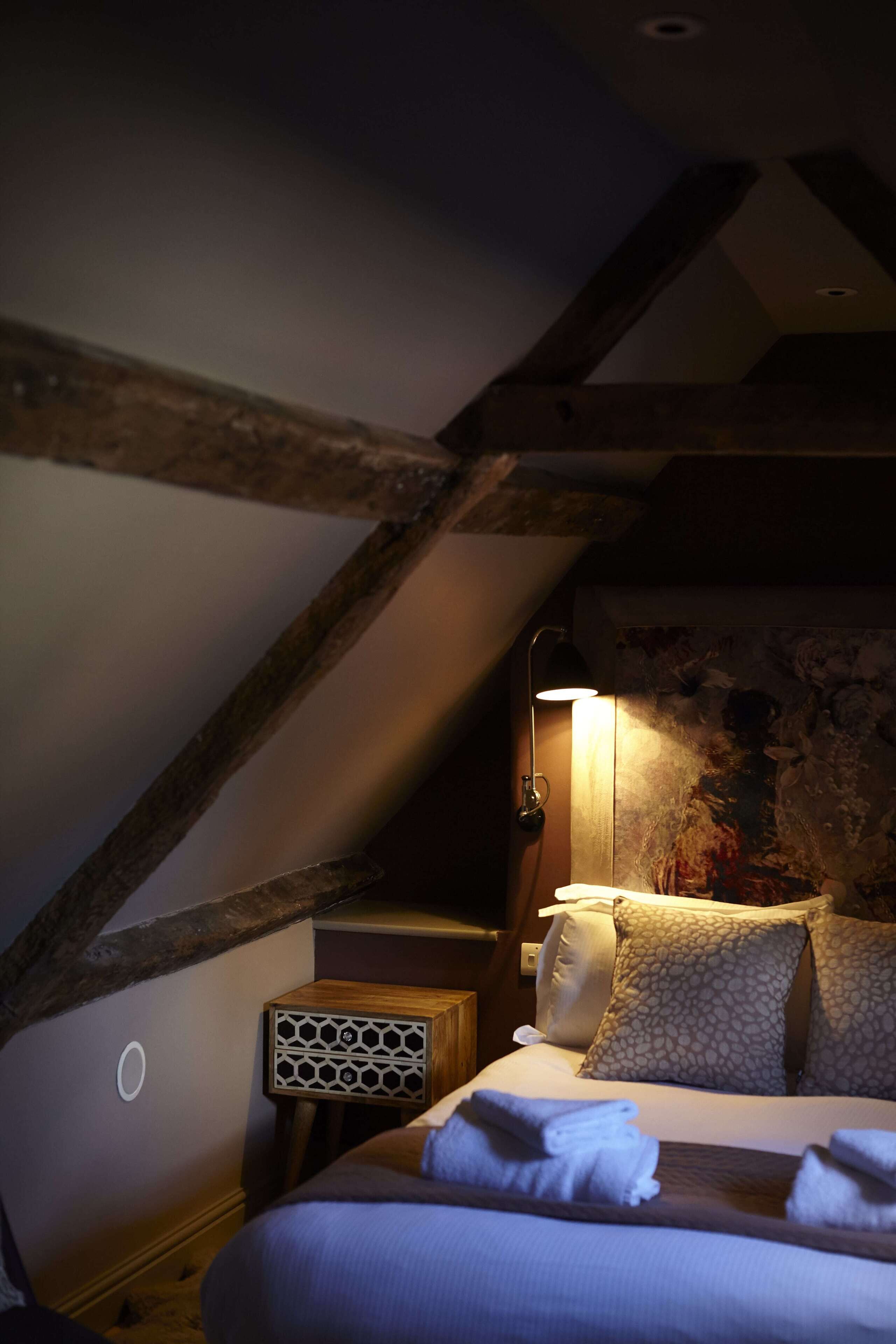 A cozy bedroom with a sloped ceiling, a bed with patterned pillows, a bedside lamp, and a small wooden nightstand with a decorative speaker.