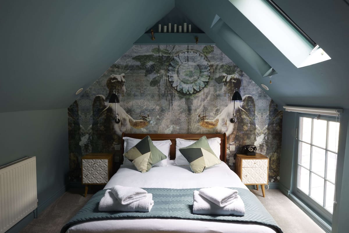 A bedroom with a sloped ceiling, a bed with two pillows, a nightstand, and a window.