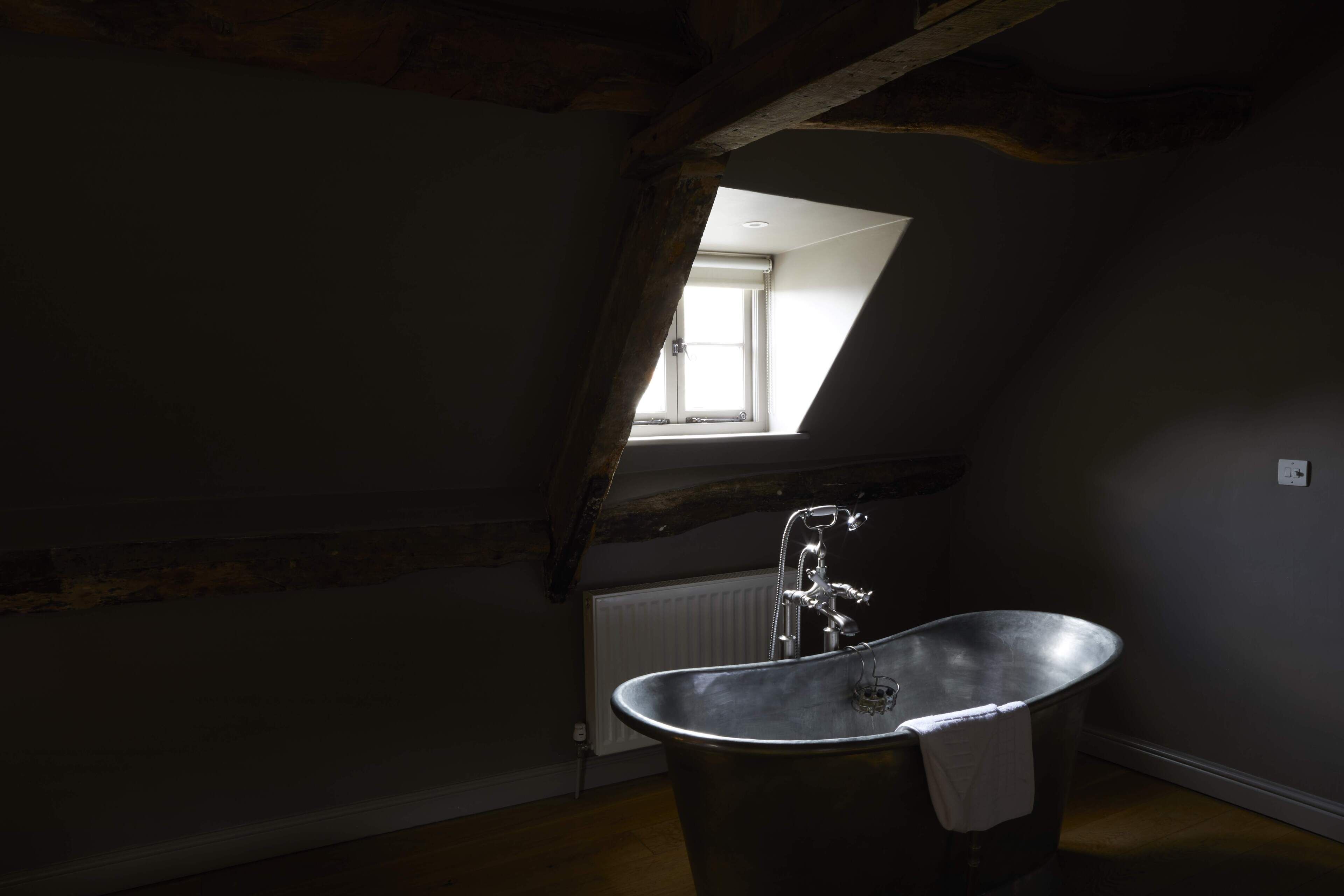 Une chambre mansardée à l’éclairage tamisé, avec un plafond en pente, une grande fenêtre et une baignoire sur pieds.