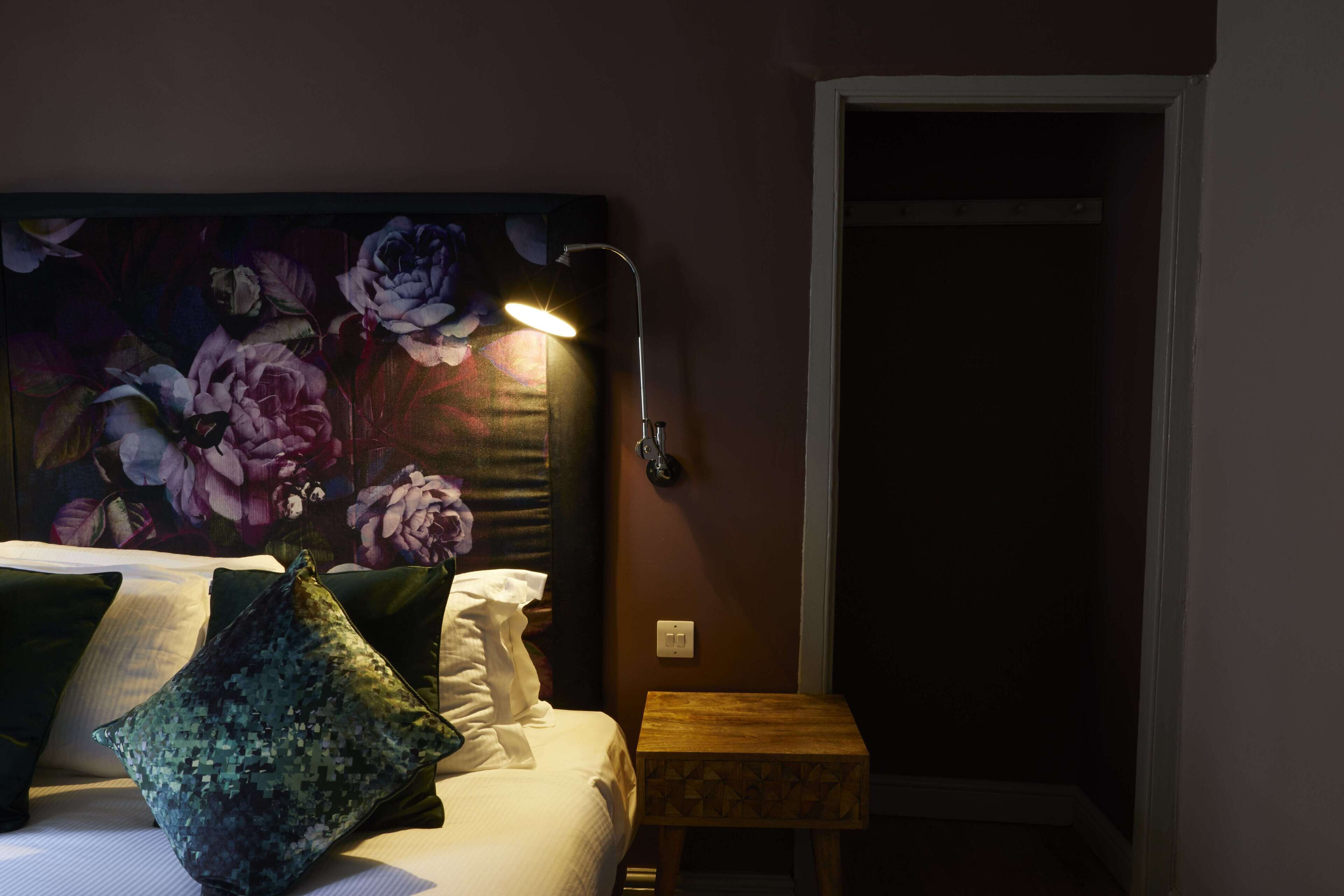 Quarto com cama, almofadas, mesa de cabeceira, luminária e cabeceira estampada com flores.