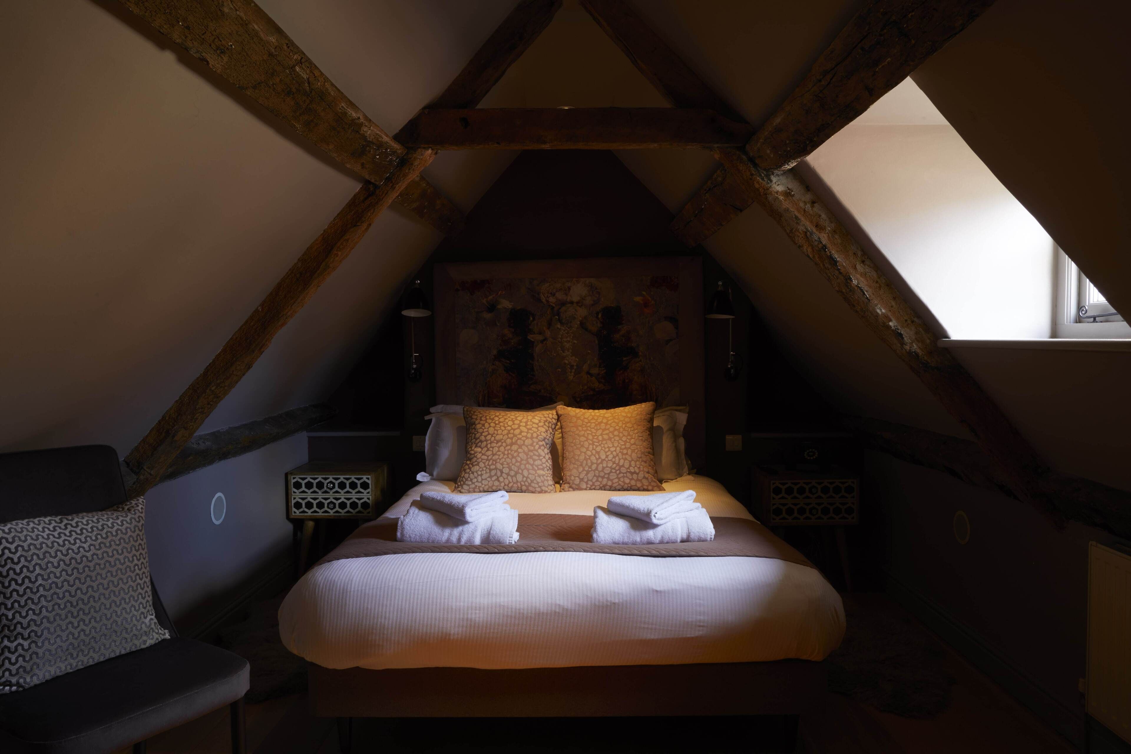 Une chambre avec un plafond en pente, un lit recouvert de draps blancs, deux oreillers, une tête de lit ornée d’un motif floral et une table de chevet avec une lampe.