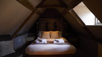 Un dormitorio con techo inclinado, cama con ropa de cama blanca, dos almohadas, cabecera con estampado floral y mesita de noche con lámpara.