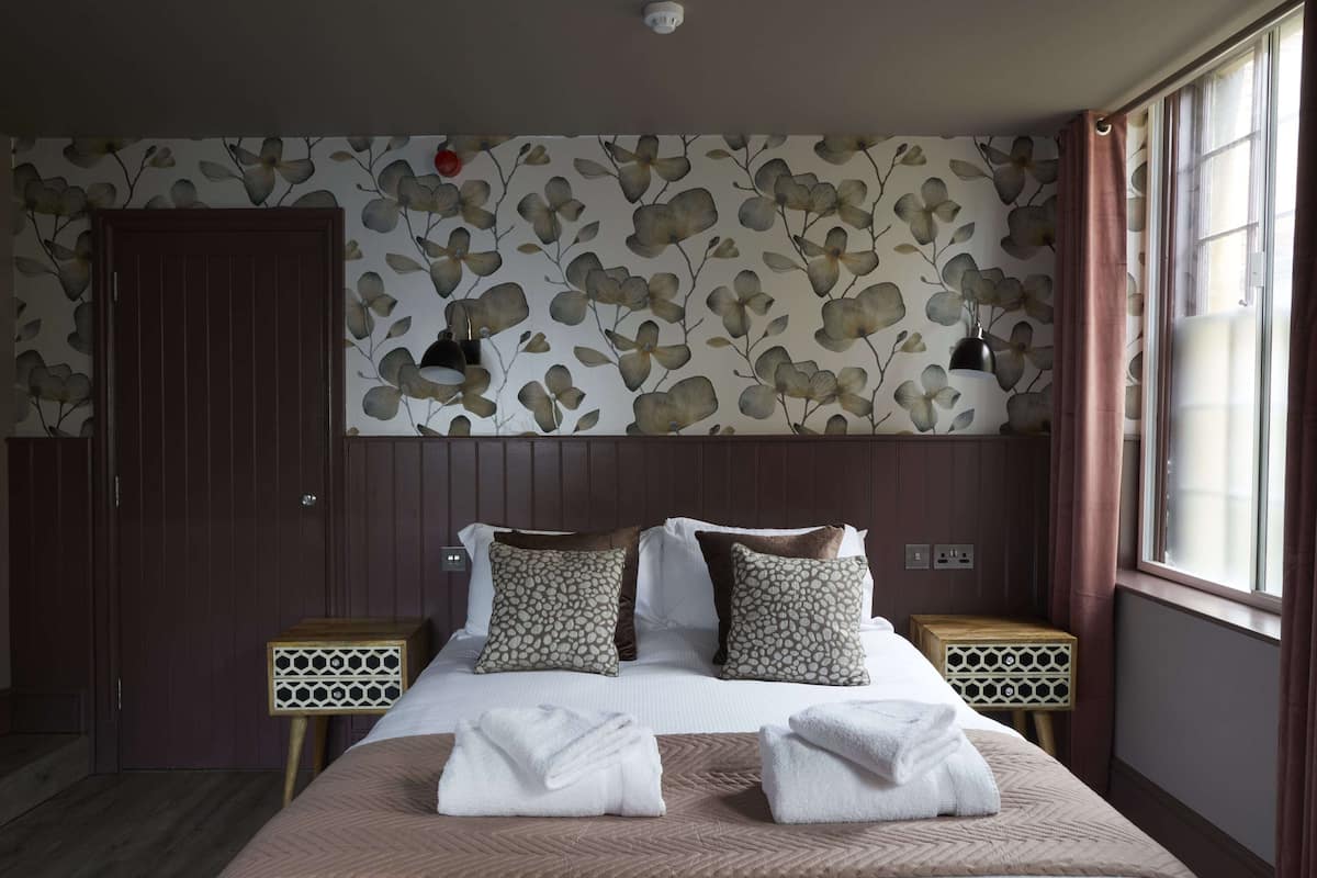 A hotel room with a bed, two bedside tables, a wall with a floral pattern, a door, and a window with curtains.